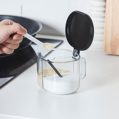 Hand pours seasoning from spoon into clear, two-compartment jar with opening lid, ideal for kitchen convenience.