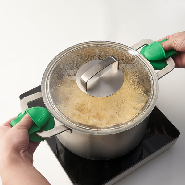 A hand holds a green silicone lid lifter, lifting a steaming metal pots lid with a metal handle on a black stove, demonstrating safe heat-resistance.