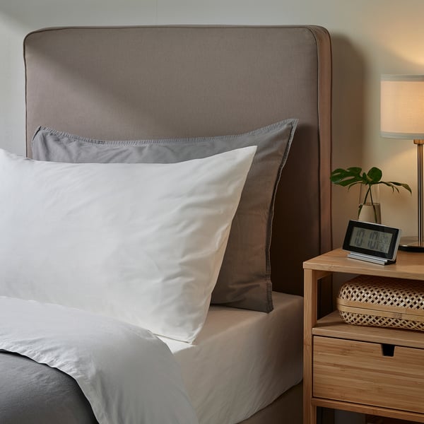 Grey pillowcase on large bed, textured headboard, bedside table with clock, lamp, plant.