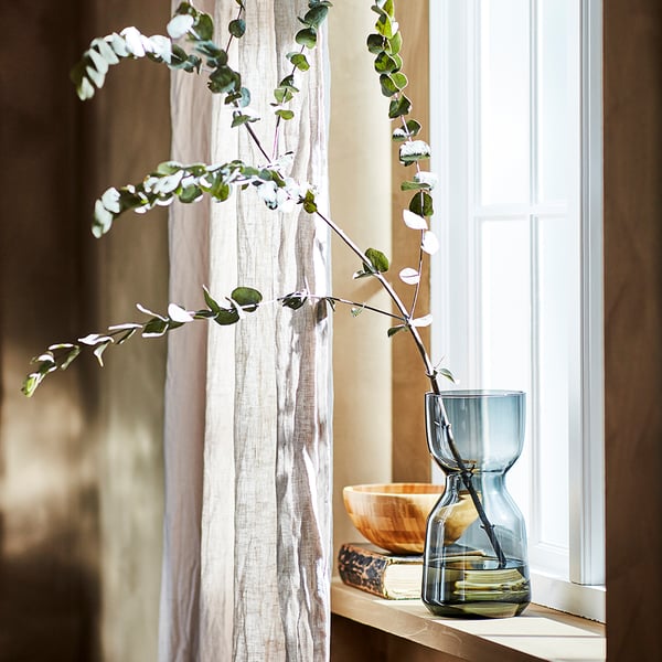 Eucalyptus branch in blue glass vase on light wood shelf, next to white linen curtains.