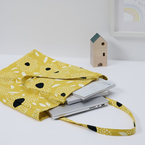Yellow tote bag with white and black patterns holding a laptop and notebooks. Small wooden birdhouse nearby.