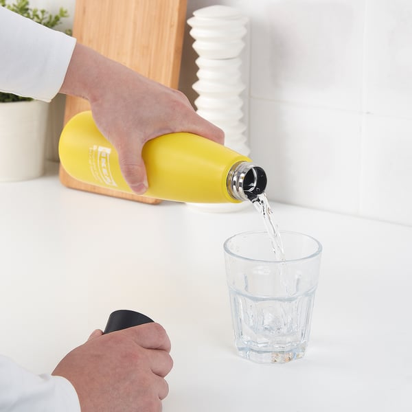 In a hand pours water from a yellow steel vacuum flask into a clear glass on a white surface, showcasing its leakproof and functional design.