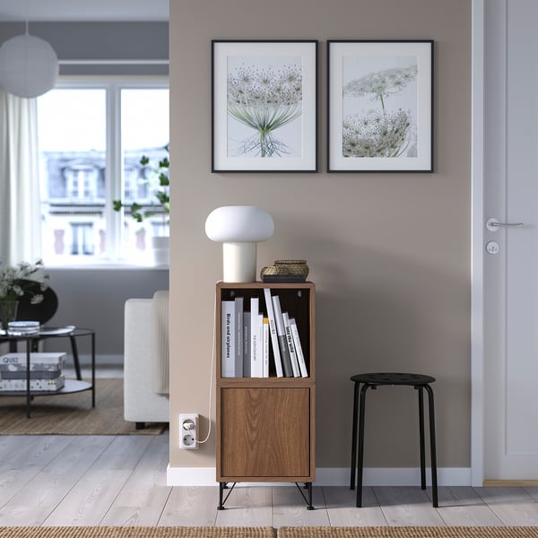 Modern living room with EKET shelf, books, lamp, stool, floral frames, and city view window.