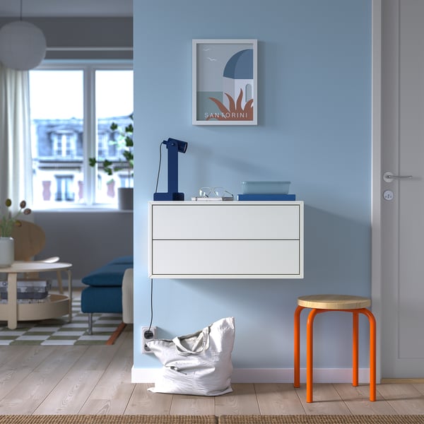 Modern living room with light blue/white decor. EKET cabinet, orange stool, santorini poster, blue lamp. Blue armchair by large window.