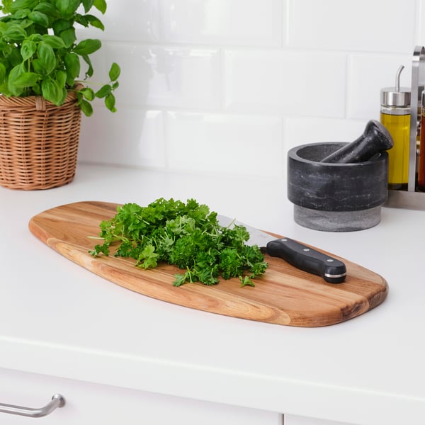 Wooden cutting board with knife and parsley on white countertop, wicker plant basket and mortar.