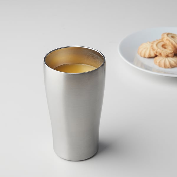 Silver FINGERVARM tumbler with golden interior, cylindrical, sits near plate of cookies.