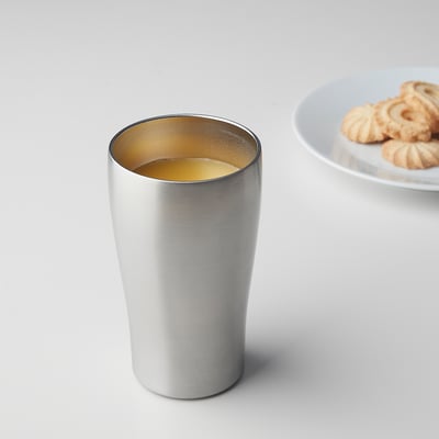 Silver FINGERVARM tumbler with golden interior, cylindrical, sits near plate of cookies.