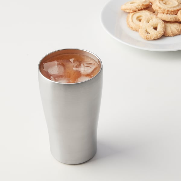 Silver stainless steel FINGERVARM cup with ice, tall and slim, sits on a table next to a plate of cookies.