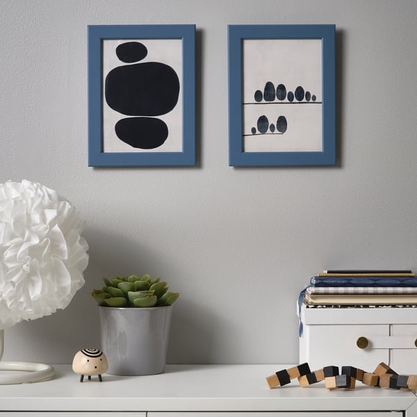 Two framed abstract artworks hang on a grey wall above a white shelf with decor items, including a plant, a wooden sheep figure, and a stack of books.