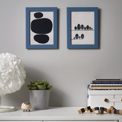 Two framed abstract artworks hang on a grey wall above a white shelf with decor items, including a plant, a wooden sheep figure, and a stack of books.