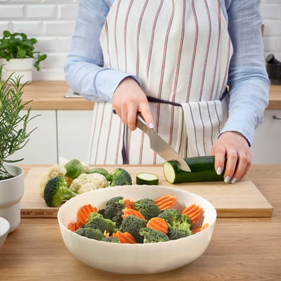 A person is slicing a cucumber on a wooden cutting board, preparing a bowl of fresh vegetables, including broccoli and carrots.