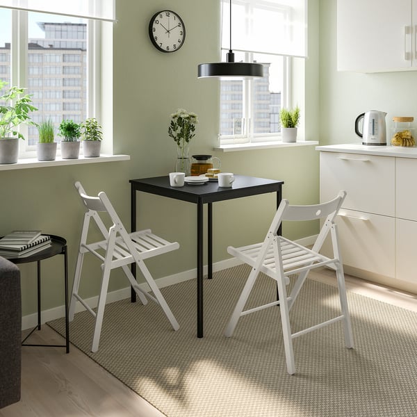 Small, modern dining setup with FRÖSVI black table & white foldable chairs. Bright, airy kitchen with green walls, large windows, & natural light.