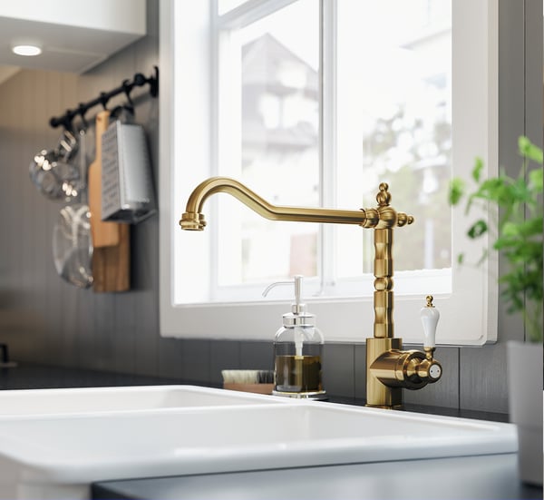 Golden brass kitchen tap with curved spout and white handles on white sink near window.