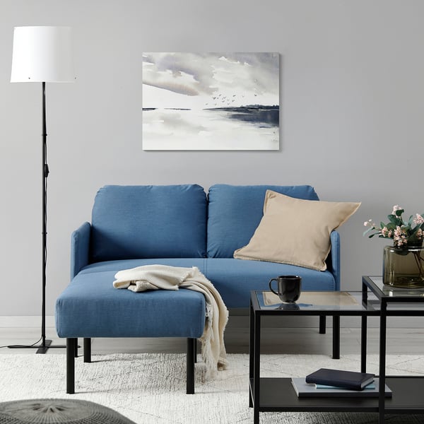 Minimalist living room: blue GLOSTAD sofa with beige pillow, black coffee table, floor lamp. Art above sofa.
