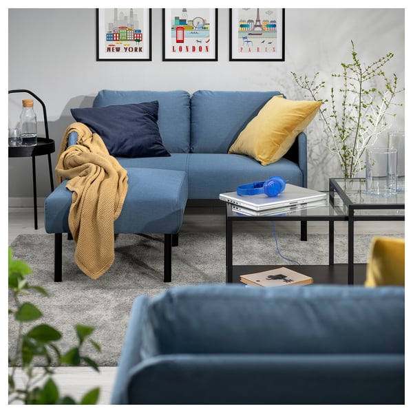 Modern living room with blue GLOSTAD couch, yellow pillows, black coffee table, blue headphones, plant, and city prints.