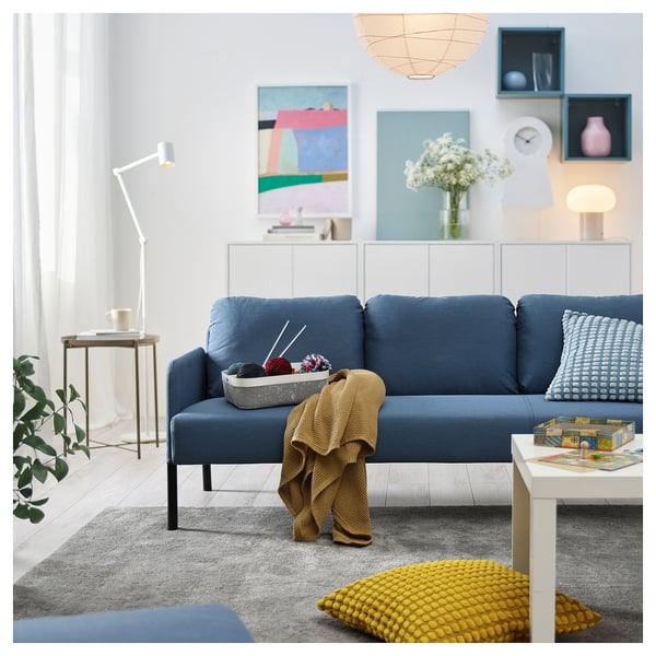 Modern living room with blue GLOSTAD sofa, yellow pillow, wooden side table, floor lamp, and decorative wall art.