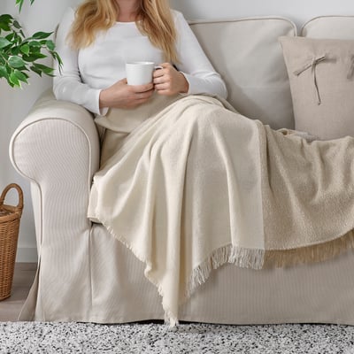 A person sits on a beige couch with a white mug, wrapped in a soft, textured throw blanket with fringes. The blanket is white and grey-beige.