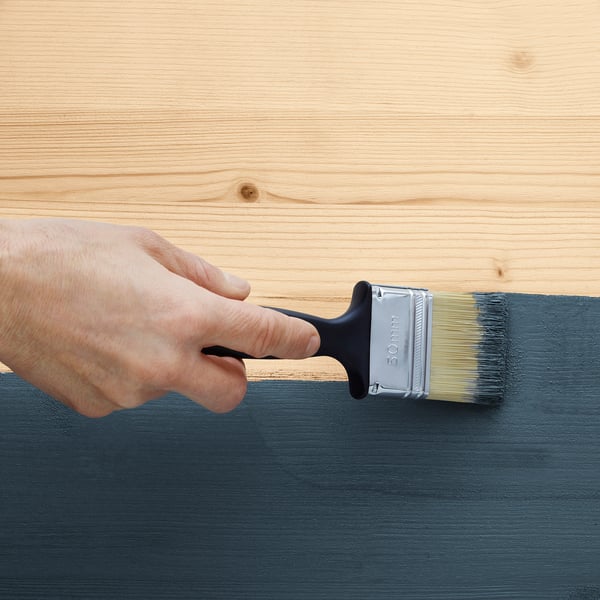 Hand holds paintbrush applying paint to wood. Brush labelled 30mm.