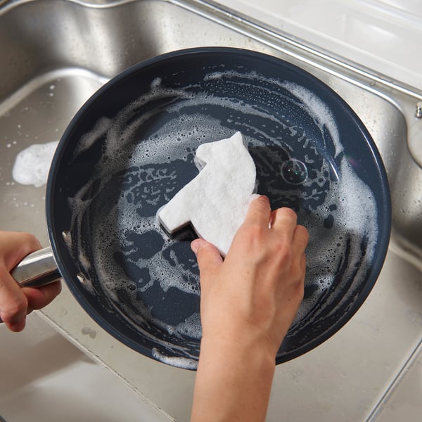 Hand cleans a black skillet with tough stains using a two-sided sponge in a sink.