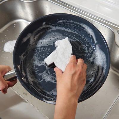 Hand cleans a black skillet with tough stains using a two-sided sponge in a sink.