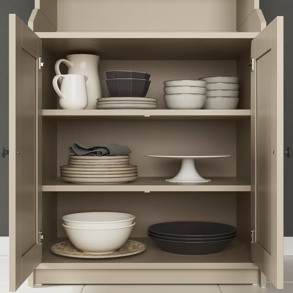 HAUGA beige cabinet with closed doors, storing white and black dishes, bowls, jug, and cake stand.