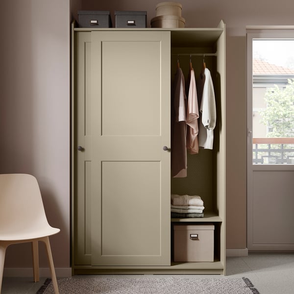 Beige HAUGA wardrobe with sliding doors, open showing clothes, boxes, and baskets. Light pink wall, white chair, modern minimalist room.