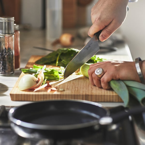 A person chops vegetables on a cutting board with a knife. A small frying pan with a non-stick coating sits nearby on a stove.