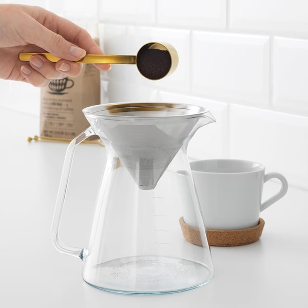 Hand pours coffee grounds into clear glass HÖGMODIG coffee maker with stainless steel philtre on white countertop.