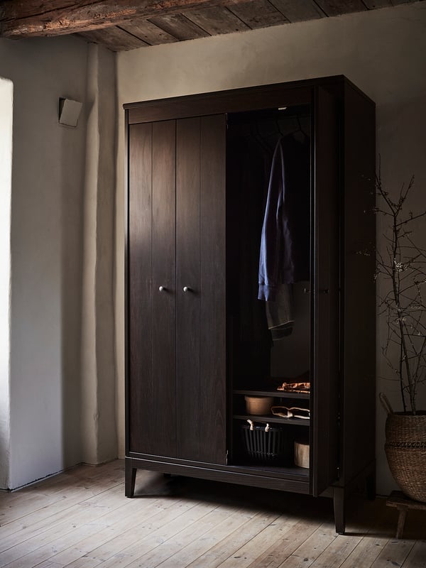 Dark wooden IDANÄS wardrobe with bifold doors, one open showing clothes and shelves. Neutral room setting.