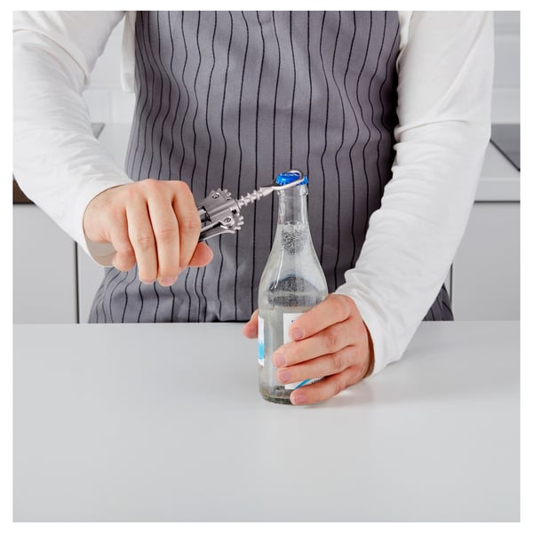 Person holding a bottle with a bottle opener on the handle, ready to open the bottle on a white surface.