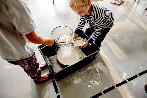 Two people prepare food at a counter using bowls and sifters with flour on a black IDEALISK tray.