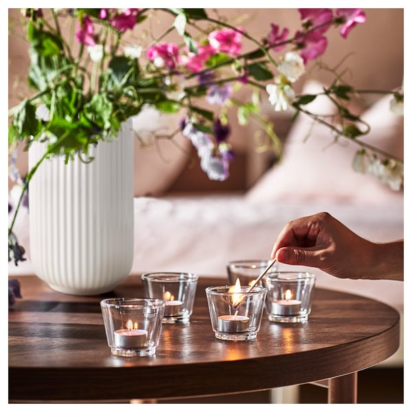 A hand lights small glass candles on a wooden table; a white vase with pink flowers sits nearby.