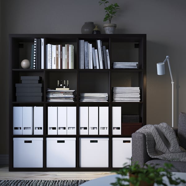 Black KALLAX shelving unit with white boxes and books, grey wall, grey armchair, grey rug, and plant.