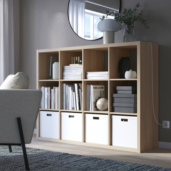 KALLAX shelving unit with books, vases, and storage boxes. Tall, with grey chair, lamp, and plants.