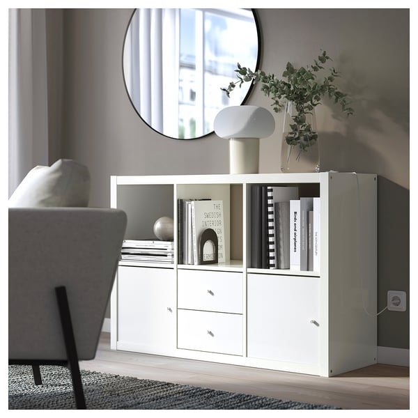 Modern living room with white KALLAX storage unit, filled with books and decorated with a lamp and plant. Grey walls and floor.