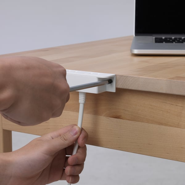 Person attaching a cord to a wooden desk, securing it with KOPPLA, a furniture charging device. Allows for easy charging of electronics.