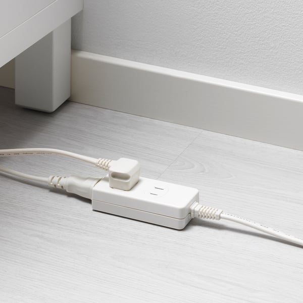 A KOPPLA extension socket lies on a light wood floor, with two white cables plugged in, extending electrical capacity; a white wall in the background.