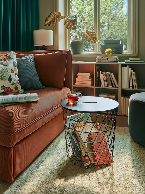 KVISTBRO red side table with books and cup, brown cushions, blue curtains, near grey rug.