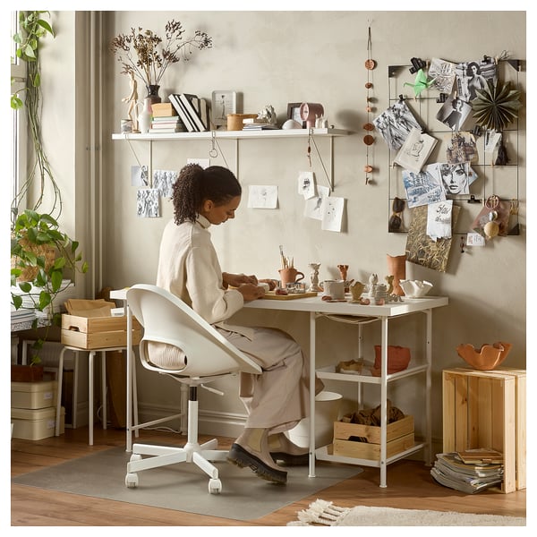 Person crafting at adjustable LAGKAPTEN desk with storage, surrounded by plants, art, and a bulletin board.
