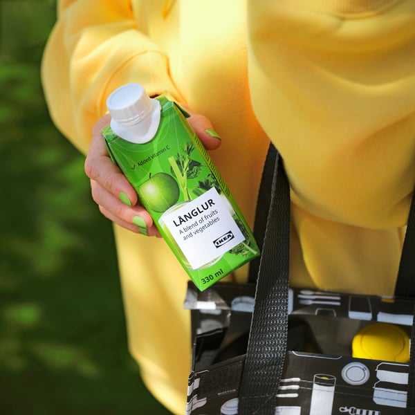Person holding a green länglur smoothie drink carton with a white cap, wearing a yellow jacket and holding a black and white reusable shopping bag.