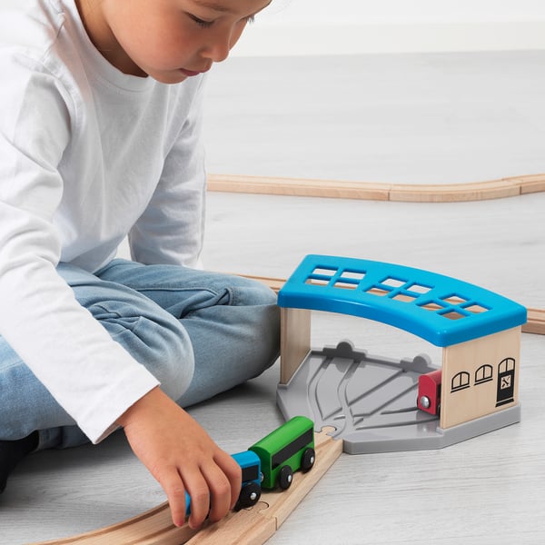 The child plays with a LILLABO wooden train set, parking a green train inside a blue and beige engine shed.