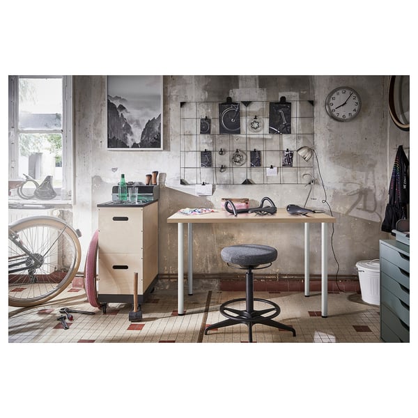 Workroom with LINNMON table, stool, tools, and artwork. Wall clock, bike, cabinets. Industrial, concrete wall, patterned floor.
