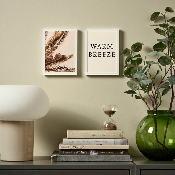 Decorated shelf with pictures, lamp, books, hourglass, and plant. Frames show palm leaf and warm breeze text in vertical and horizontal orientations.