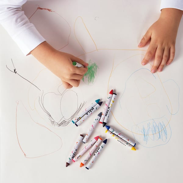 Childs hands drawing with colourful crayons. Several MÅLA crayons are scattered on the paper, which abstract drawings in various colours.