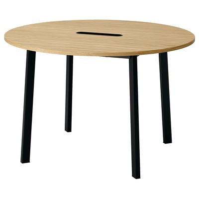 A modern round table named MITTZON. It has a light wooden top and black metal legs. Designed for meetings, it a cable management tray.