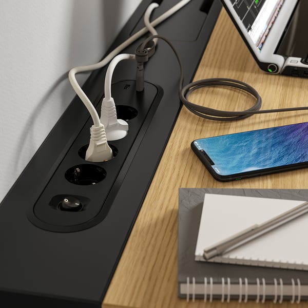 A wooden desk with a cable management tray and built-in power sockets, featuring a smartphone, notebook, and headphones.