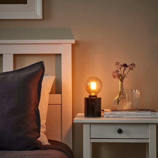Bedroom bedside table with lit amber MOLNART bulb, vase, jars, magazines, & bed with peach pillows.