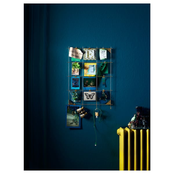 A teal wall displays a brass-plated wire grid with various pictures and notes clipped in. Below, a bright yellow radiator is present.