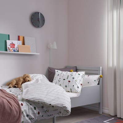 Bedroom with NATTSLÄNDA duvet on twin bed, grey clock, books, lamp, purple pillows, teddy bear, curtain.
