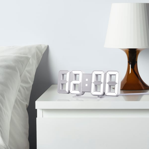 NOLLÅTTA alarm clock on bedside table, four digital displays, sleek minimalist look with lamp.